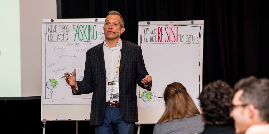 Curtis Michelson delivering a change-leadership talk on stage, gesturing between two hand-lettered flip charts as the audience looks on